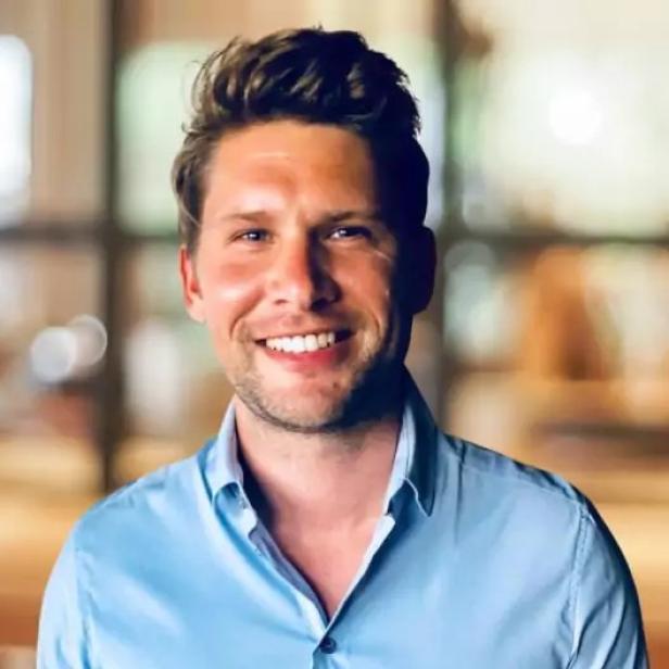 Mann mit blonden Haaren und blauem Hemd lächelt vor unscharfem Hintergrund.