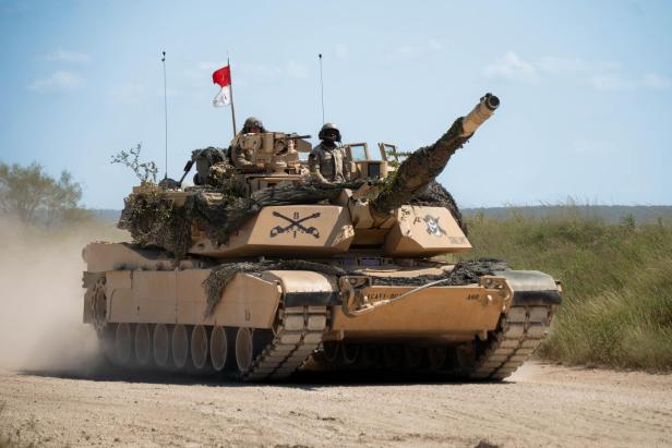 Kampfpanzer mit Tarnnetz und Soldaten fährt auf staubiger Piste durch offene Landschaft.