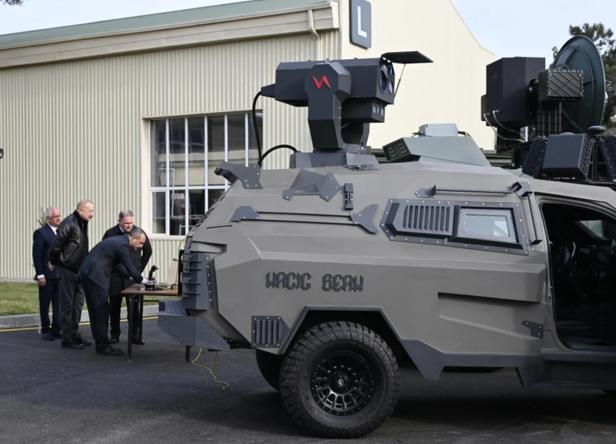 Gepanzertes Fahrzeug mit Laserwaffe Magic Beam, daneben mehrere Männer im Anzug und Lederjacke.