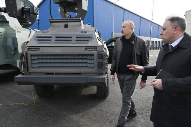 Zwei Männer begutachten ein gepanzertes Militärfahrzeug vor einer blauen Industriehalle.