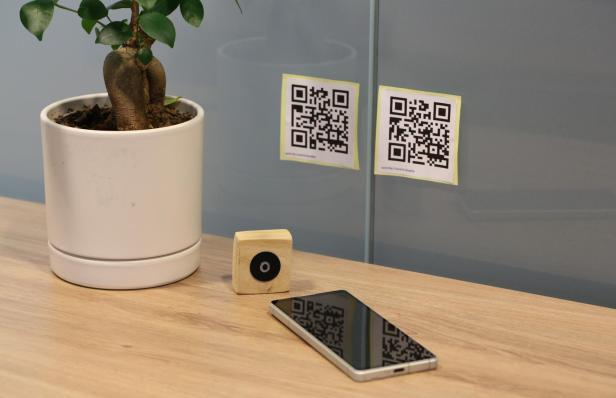 zwei QR-Codes an einer Glaswand, davor eine Zimmerpflanze, der NFC-Holzklotz und ein Smartphone