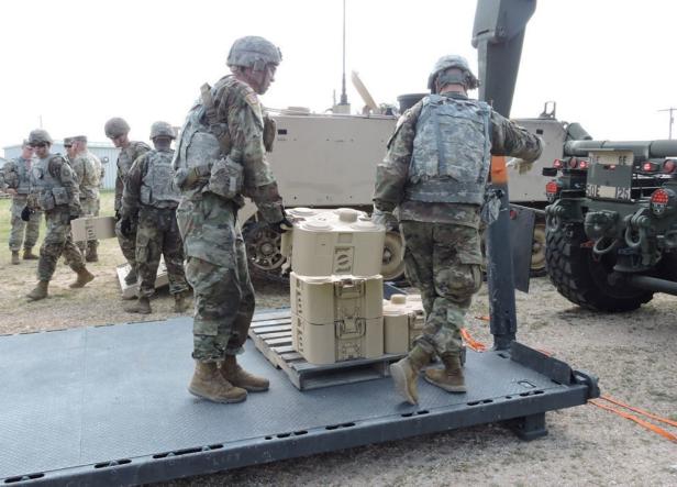 Soldaten in Tarnuniform verladen beige XM204-Systeme auf eine Palette neben Militärfahrzeugen.