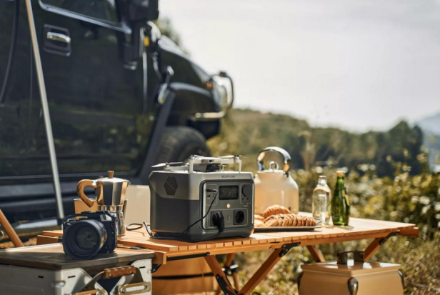 Powerstation auf Campingtisch mit Kamera, Kaffeekanne, Brot und Getränken im Freien.