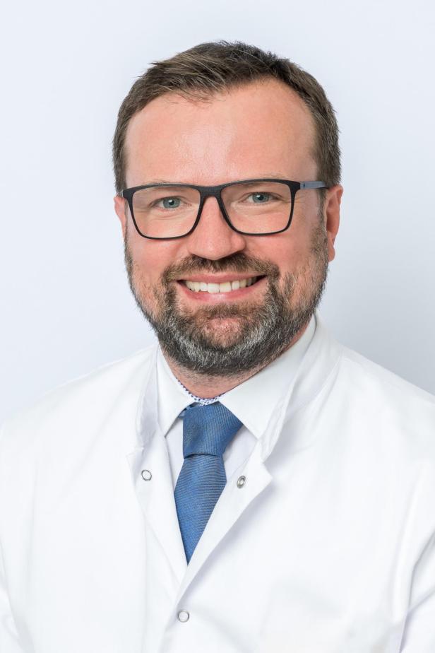 Mann mit Brille, Bart und blauer Krawatte trägt einen weißen Arztkittel und lächelt freundlich.