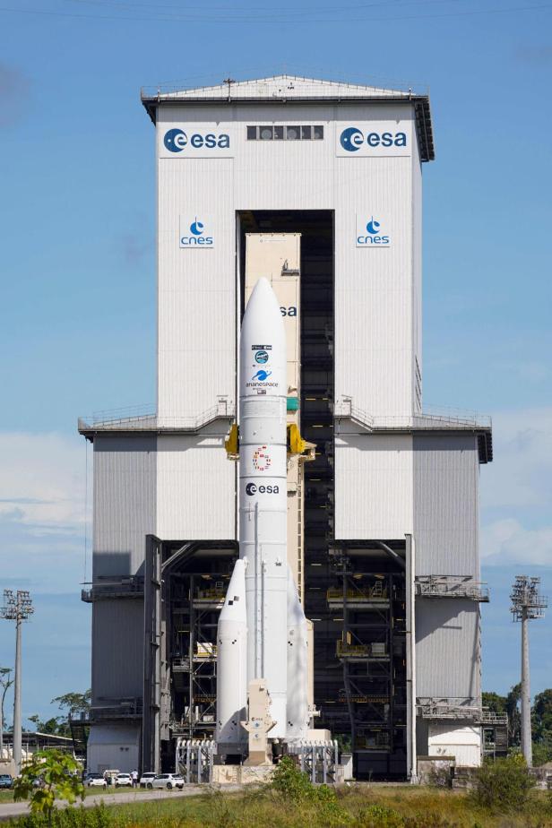 Eine große weiße Rakete steht vor einem Gebäude mit ESA- und CNES-Logos auf einer Startrampe.