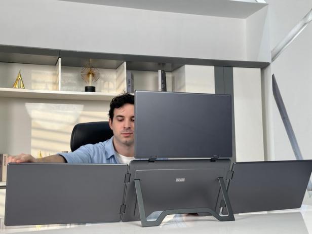 Mann sitzt am Schreibtisch hinter einem Laptop mit drei zusätzlichen ausklappbaren Bildschirmen.