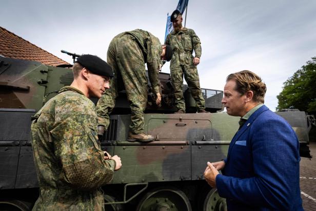 Mehrere Soldaten in Tarnuniform stehen auf einem Militärfahrzeug, während ein Mann im blauen Anzug mit ihnen spricht.
