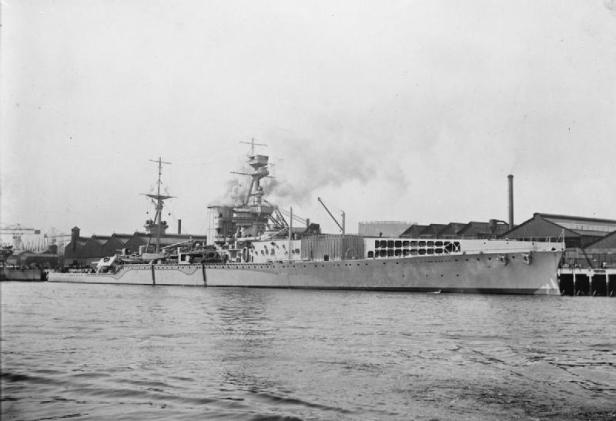 Ein Kriegsschiff mit Flugdeck liegt an einem Kai vor Werftgebäuden.