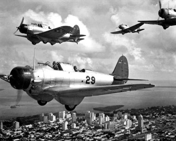 Vier Militärflugzeuge fliegen in Formation über eine Stadtlandschaft mit Wasser im Hintergrund.