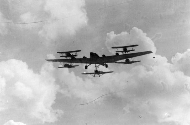 Ein großes Flugzeug mit mehreren kleinen Doppeldeckern darunter fliegt vor bewölktem Himmel.