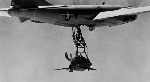 Ein großes Flugzeug lässt ein kleines Jagdflugzeug an einem Trapez aus dem Rumpf herab.