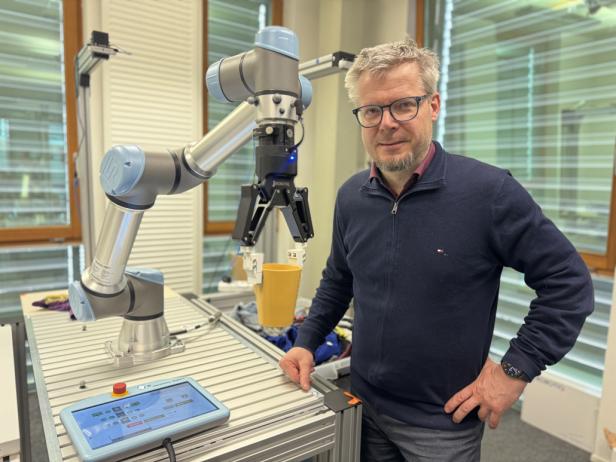 Ein Mann steht neben einem Roboterarm, der einen gelben Becher mit Greifern hält.