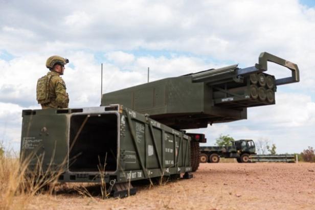 Soldat steht neben einem HIMARS-Raketenwerfer auf einem Feld unter bewölktem Himmel.