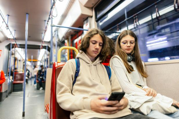 Zwei Jugendliche sitzen nebeneinander in einer Straßenbahn, einer schaut auf sein Handy, die andere blickt nach unten.