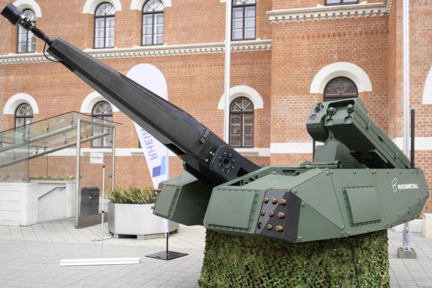 Grünes, modernes Waffensystem mit langem Geschützrohr und Raketenwerfer vor einem Backsteingebäude.