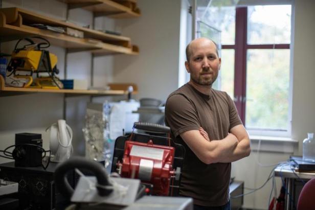Mann mit verschränkten Armen steht in einem Laborraum mit technischen Geräten und Regalen.