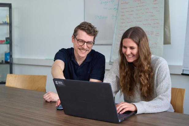 Zwei Personen arbeiten gemeinsam lächelnd an einem Laptop in einem Besprechungsraum.
