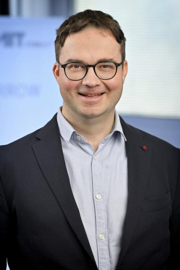 Mann mit Brille und dunklem Sakko lächelt, trägt ein helles Hemd, unscharfer Hintergrund mit Blautönen.