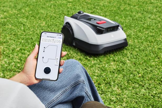 Eine Person steuert einen Mähroboter per Smartphone-App auf einer grünen Rasenfläche.