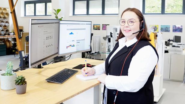 Frau mit Headset berät an Schreibtisch mit zwei Monitoren im modernen Büro.