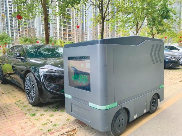 Lade-Roboter steht neben einem schwarzen Elektroauto auf einem Parkplatz in einer Wohnsiedlung.