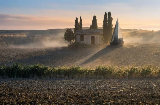 Eine moderne, schwarze Pyramide steht neben einem alten Häuschen auf einem Feld bei Sonnenuntergang.