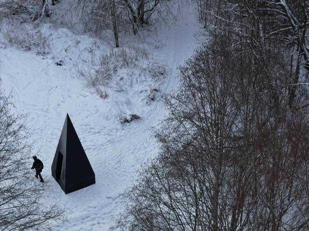 Eine schwarze, pyramidenförmige Hütte von The Klumpen steht im verschneiten Wald, eine Person nähert sich dem Eingang.