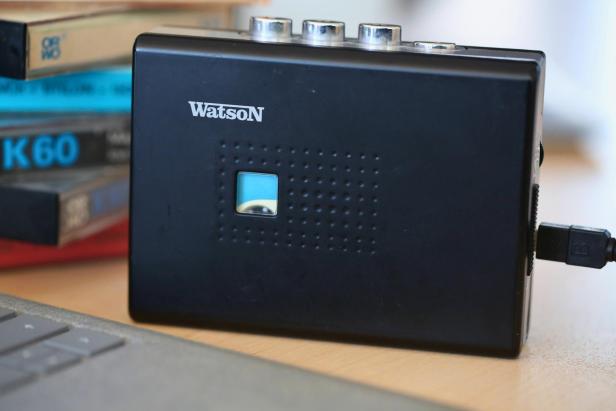 Ein Kassettenspieler der Marke Watson ist per USB-Kabel mit einem Computer verbunden, im Hintergrund liegen mehrere alte Musikkassetten.