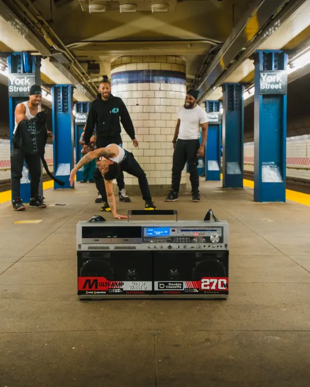 Vier junge Männer tanzen und lachen in einer U-Bahn-Station, vorne steht eine große Boombox.