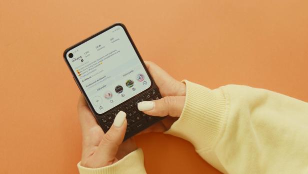Person hält ein Smartphone mit physischer Tastatur vor orangefarbenem Hintergrund.