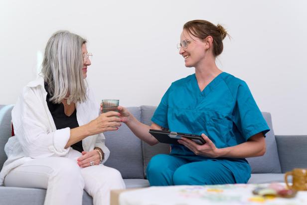 Pflegekraft reicht einer älteren Frau ein Glas Wasser und hält ein Tablet in der Hand.