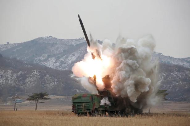 Ein nordkoreanischer Mehrfachraketenwerfer feuert eine Rakete in einer ländlichen, bergigen Landschaft ab.