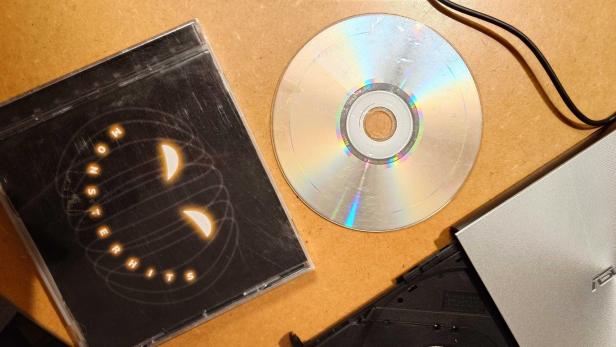 Zerkratzte CD, Hülle mit „Monsterhits“-Cover und externes CD-Laufwerk auf Holztisch.