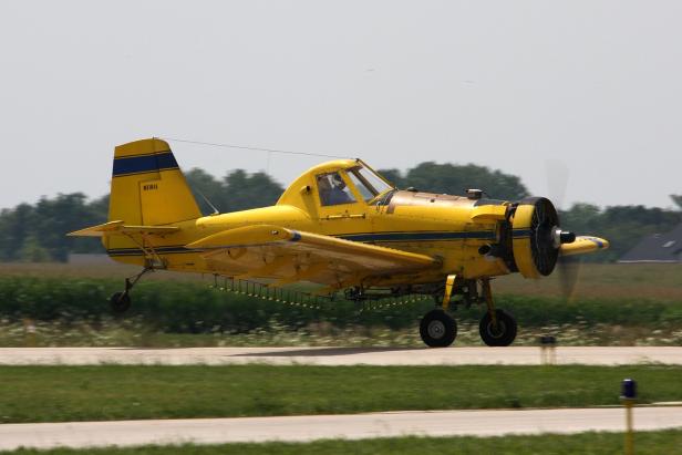 Gelbes Sprühflugzeug beim Starten auf einer Landebahn mit grünem Feld im Hintergrund.