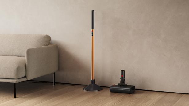 Ein schlanker, kupferfarbener Nassreiniger steht neben einer Ladestation und einem Sofa auf Holzboden.
