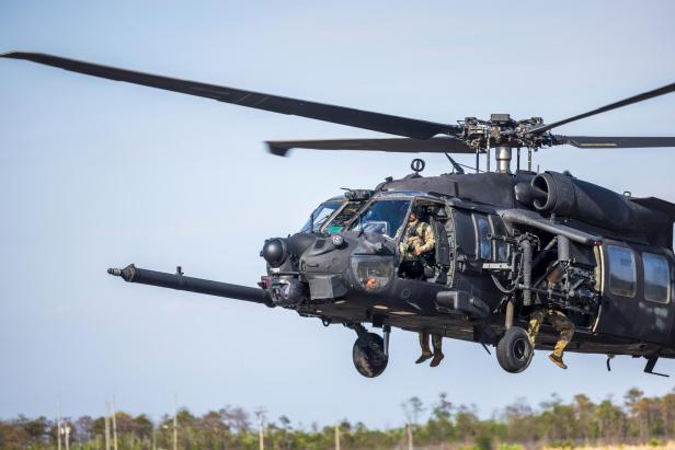 Schwer bewaffneter Black Hawk Hubschrauber mit Soldaten im Flug über offenem Gelände.