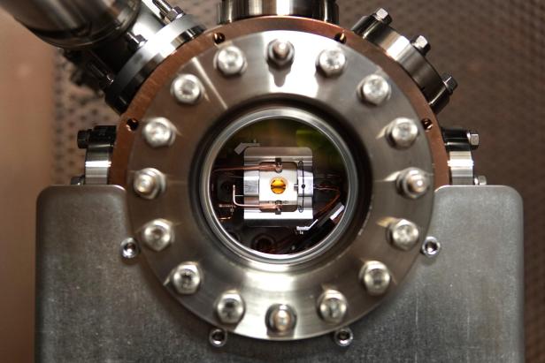 Rundes Metallgehäuse mit Schrauben und Sichtfenster, dahinter technische Bauteile und ein gelber Punkt in der Mitte.