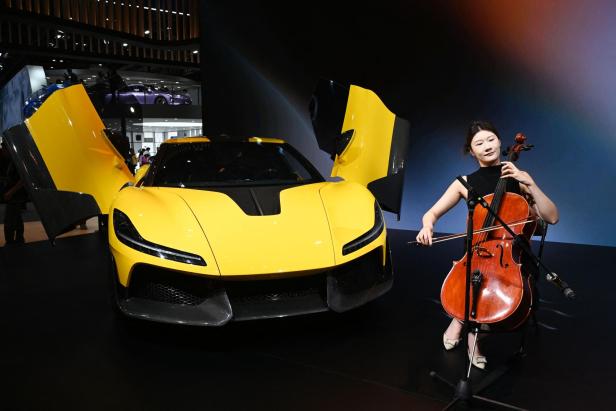 Eine Frau spielt Cello neben einem gelben Sportwagen mit geöffneten Flügeltüren auf einer Ausstellungsfläche.
