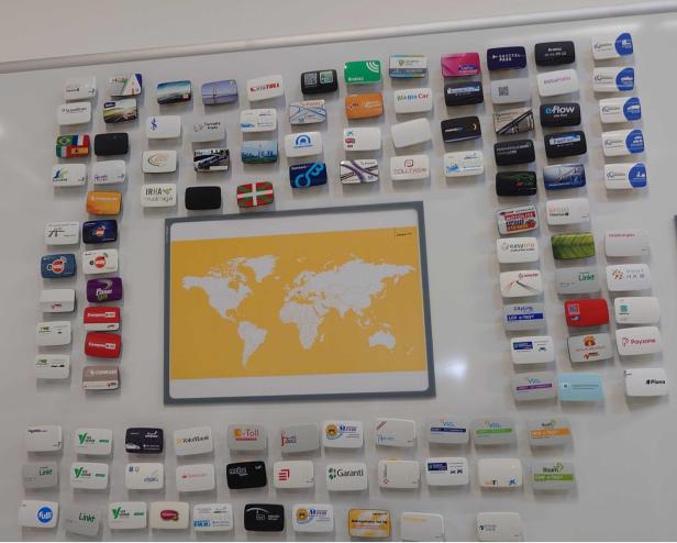 Verschiedene Mautgeräte aus aller Welt sind rund um eine Weltkarte an einer Wand angebracht.