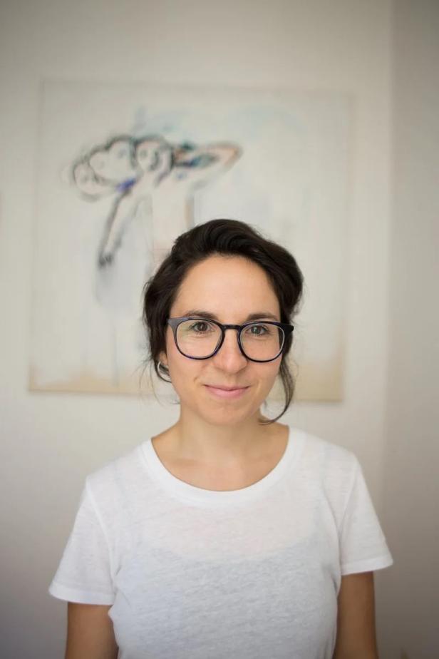 Frau mit dunklen Haaren und Brille trägt ein weißes T-Shirt und lächelt vor einem unscharfen Gemälde im Hintergrund.
