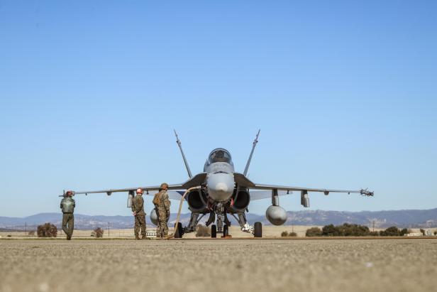 Kampfjet steht auf Rollfeld, drei Soldaten in Uniform und Helm daneben, Berge im Hintergrund.