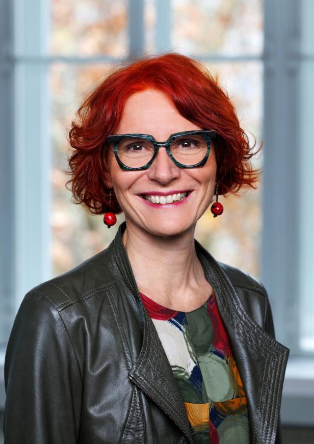 Frau mit roten Haaren, großer Brille, buntem Oberteil und Lederjacke lächelt vor einem Fenster mit unscharfem Hintergrund.