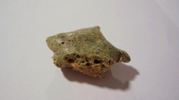 Poröses, grünlich-graues Stück Trinitit auf hellem Untergrund.