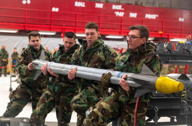 Vier Soldaten in Tarnuniform heben gemeinsam eine große Luft-Luft-Rakete mit gelber Spitze.