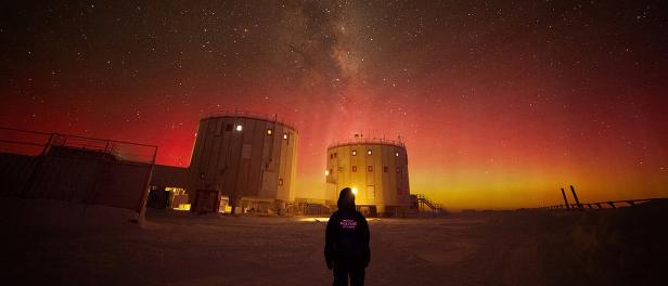 Eine Person steht vor der Antarktisstation Concordia, umgeben von Schnee und beleuchtetem Nachthimmel mit Polarlichtern.