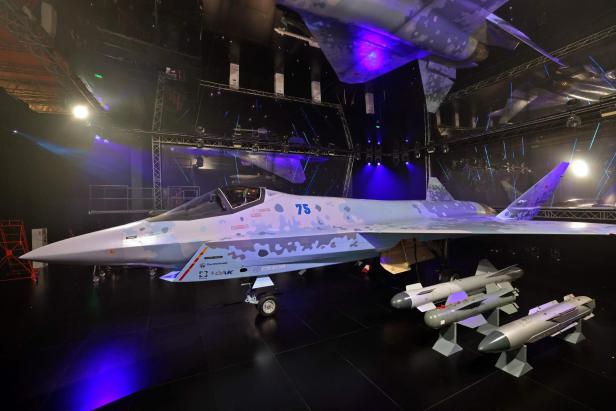 Ein modernes Tarnkappen-Kampfflugzeug mit hellblauer Tarnung steht in einer Halle, daneben liegen mehrere Raketen auf Ständern.
