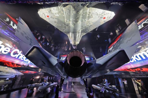 Ein modernes Kampfflugzeug mit Tarnmuster steht in einer Halle, umgeben von Menschen und Raketen.