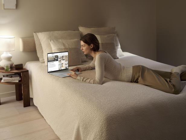 Eine Person arbeitet entspannt mit einem Notebook auf einem Bett in einem gemütlichen Schlafzimmer.