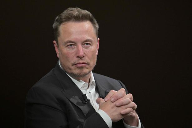 Elon Musk trägt einen dunklen Anzug mit weißem Hemd und blickt mit gefalteten Händen ernst in die Kamera vor einem dunklen Hintergrund.