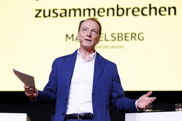 Ein Mann im blauen Sakko spricht mit Mikrofon und Papier in der Hand vor einer gelben Präsentationsfolie.
